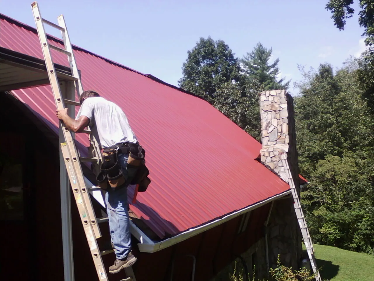 Roofer on a ladder beside a red metal roof and stone chimney — Soffit & Fascia Repair by Roofing Brothers in Washington Court House